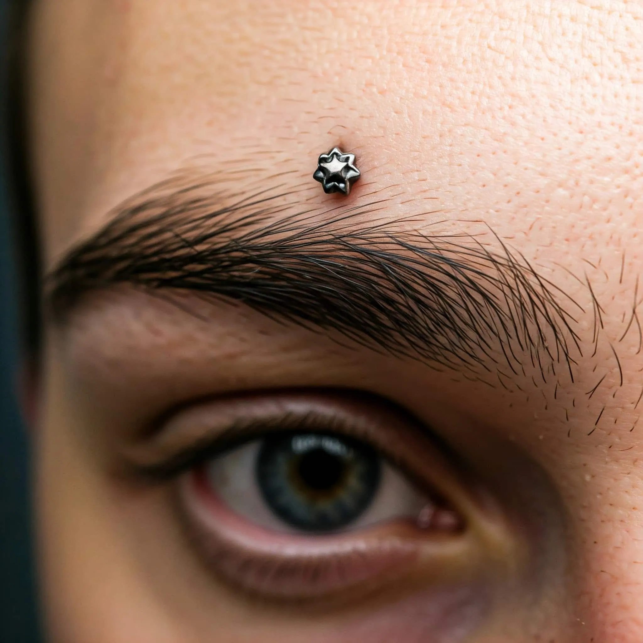 A detailed view of a man's face featuring a surface eyebrow piercing on his forehead, emphasizing his striking features.