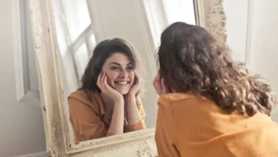 A young woman with curly brown hair and a mustard-colored top smiles warmly at herself in an ornate mirror.